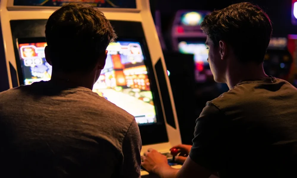 Two friends playing a retro fighting game arcade cabinet in a dimly lit games arcade, screen glow on their faces.