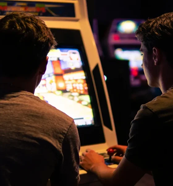Two friends playing a retro fighting game arcade cabinet in a dimly lit games arcade, screen glow on their faces.