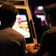 Two friends playing a retro fighting game arcade cabinet in a dimly lit games arcade, screen glow on their faces.