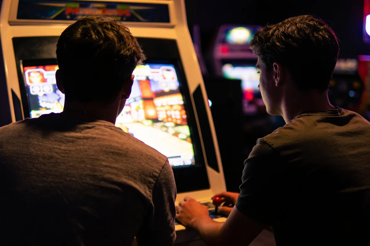 Two friends playing a retro fighting game arcade cabinet in a dimly lit games arcade, screen glow on their faces.