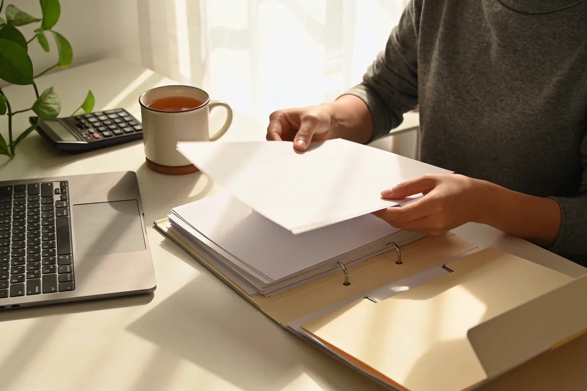 A person sorting blank papers into folders at a tidy home office desk in natural light.