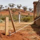 A short timber survey peg with a bright nail in its centre standing in red-brown earth beside a leaning stake, with a taut white string line running to the corner of an older brick fence and a surveyor's tripod out of focus beyond