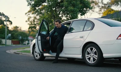 A passenger stepping into the back seat of a waiting car on a quiet suburban street in daylight.