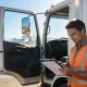 An owner driver in a hi-vis vest reviewing paperwork beside an open truck door in a truck yard.