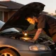 A mechanic checking a car engine in a suburban driveway in the late afternoon sun.