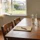 A timber dining table in an ordinary Australian home set for a meeting that has not started, with a glass jug and three glasses, a closed manila folder and pen and a potted plant, morning light coming through a curtained window onto a timber floor