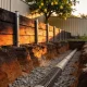 A domestic retaining wall of dark timber sleepers held by steel posts seen from the excavated side, with an open trench of coarse gravel and a slotted drainage pipe along its base and a spade standing in the spoil beside a lemon tree above