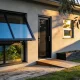 The rear wall of a modern Australian home in late-afternoon light, with a large black-framed awning window standing open outward from the bottom beside a black-framed door and a smaller window, above a timber deck and planter box with a gum tree in the foreground