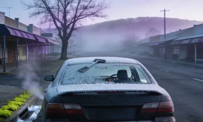 A dark sedan standing at the kerb of an empty country town street on a hard winter morning, its roof and rear window thickly frosted with an ice scraper lying on the boot and vapour drifting behind, shopfront awnings along the footpath and low hills disappearing into fog at the end of the street