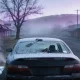 A dark sedan standing at the kerb of an empty country town street on a hard winter morning, its roof and rear window thickly frosted with an ice scraper lying on the boot and vapour drifting behind, shopfront awnings along the footpath and low hills disappearing into fog at the end of the street